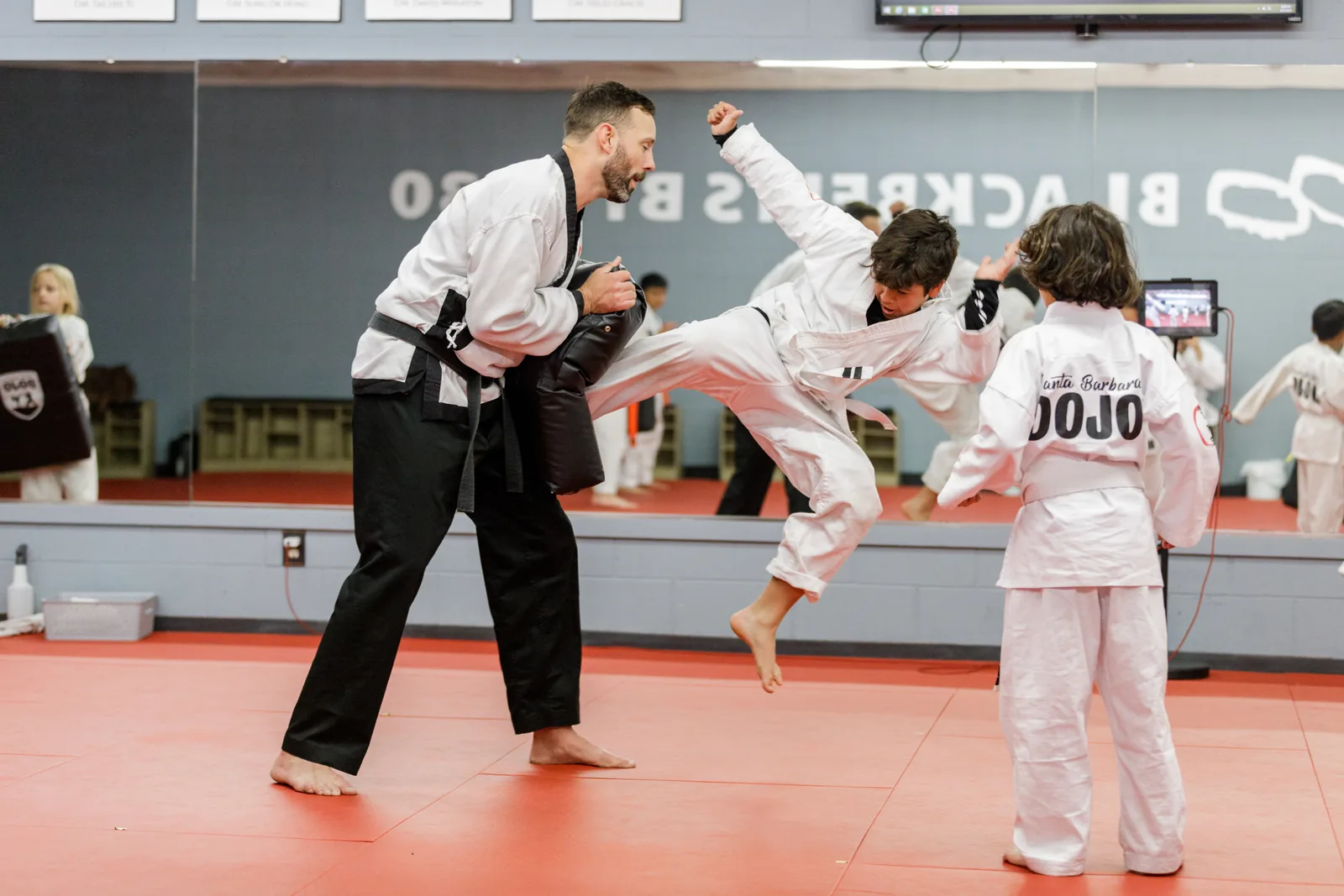 An instructor guides a child's kick while classmates watch, building confidence at Santa Barbara Dojo