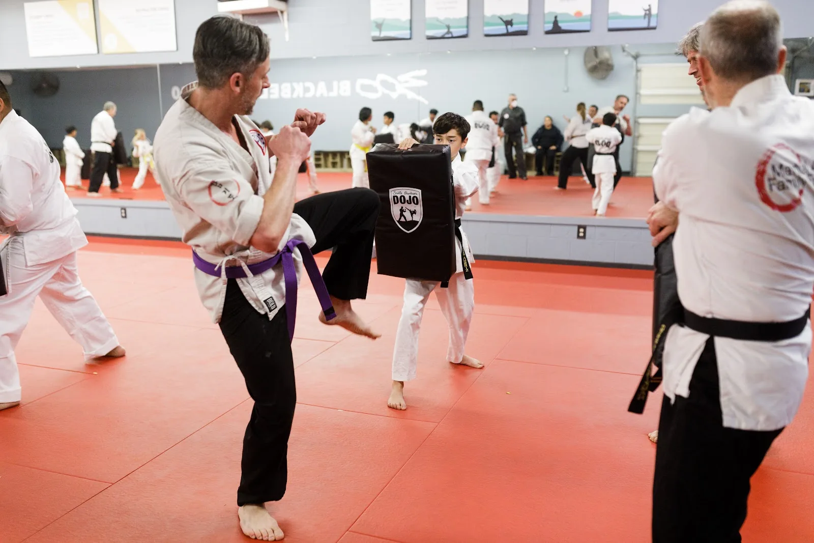 A parent and child in uniform practice a kick together while an instructor watches at Santa Barbara Dojo